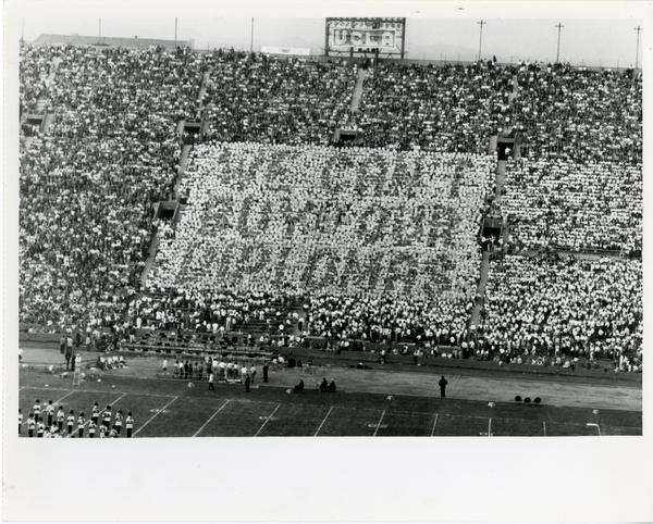 Crowd holds cards that say "We Can't Buy Our Diplomas" at the football game, ca. 1950
