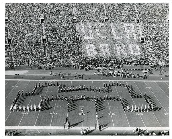 Marching band forms the shape of Royce Hall on footbal field with crowd holding up signs that spell "UCLA BAND" in the background, 1971