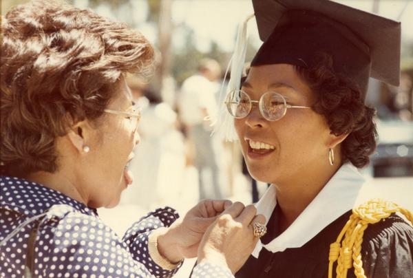 Woman having her robe adjusted by a woman, possibly her mother, at commencement, 1974