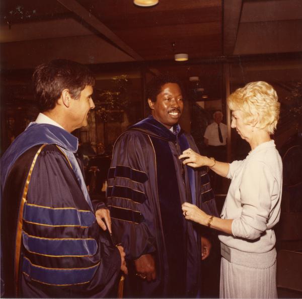 Woman assists two platform party members with their robes for commencement, June 1979