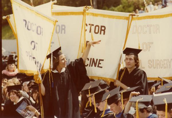 Graduate holding a department banner waves to those in the audience at commencement, June 1979