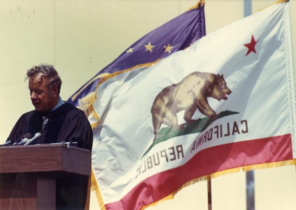 Member of the party platform addresses the audience with the California flag flying behind him at commencement