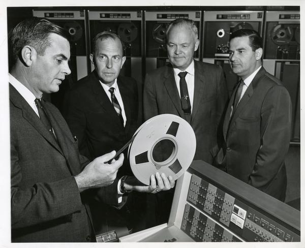 One worker holds a reel of taype while three others, including chancellor Franklin D. Murphy, look on at the Western Data Processing Center, ca. 1960's