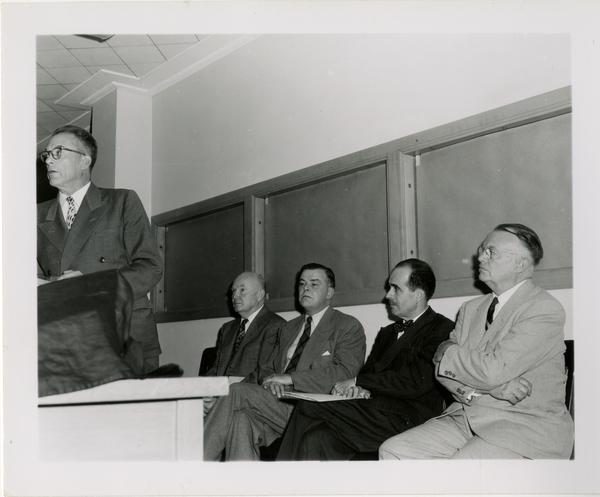 Looking at men including Lawrence Clark Powell, sitting behind speaker at podium during Friends of the UCLA Library event