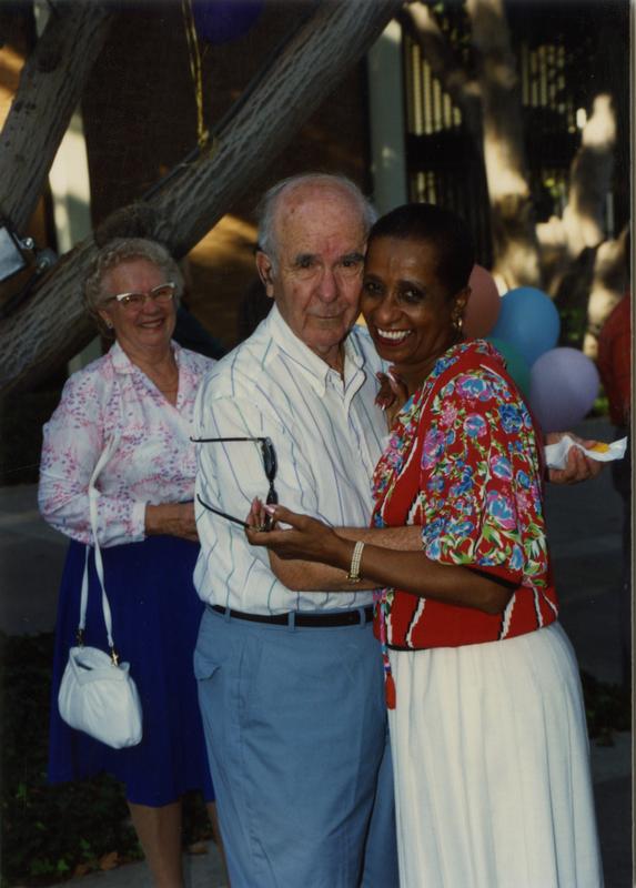 Library staff embrace each other for the camera with another staff member smiling in the background at a staff retirement party, 1991