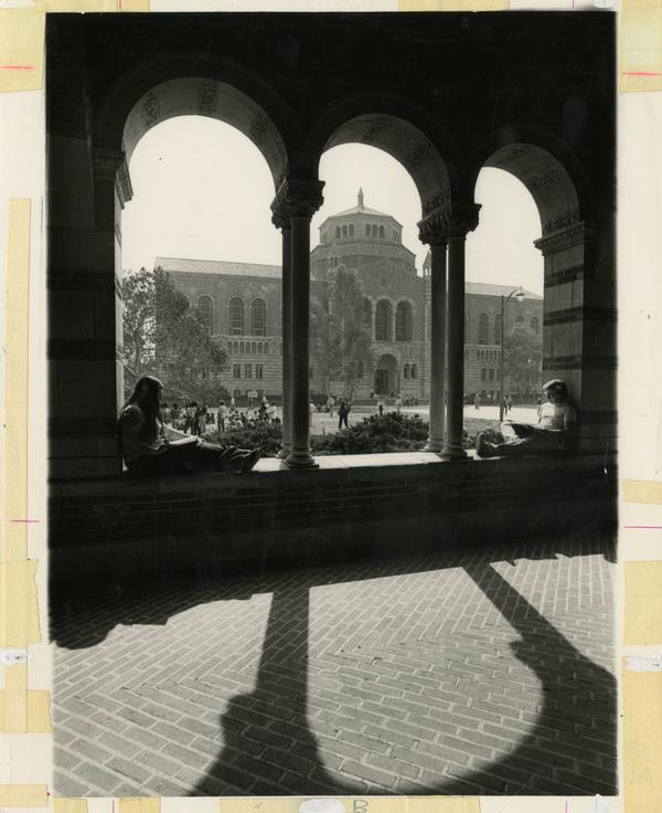 Students sitting in Royce Hall arches with Powell Library in background