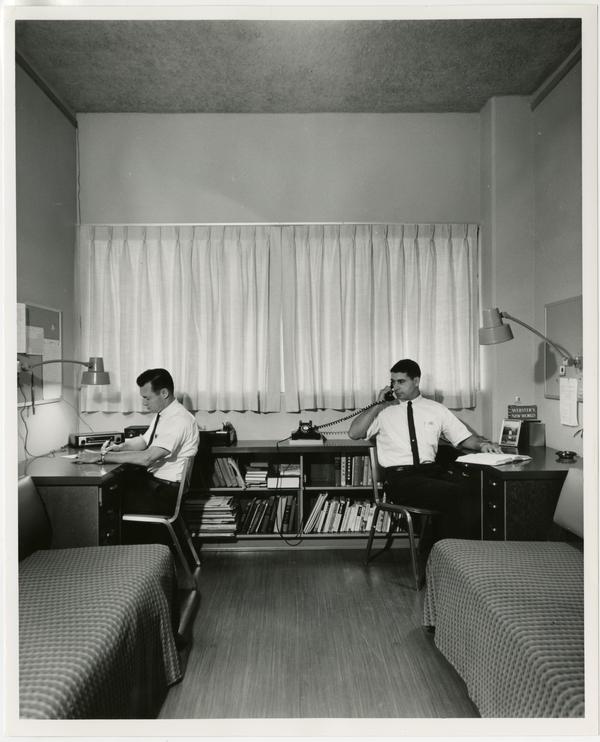 Interior view of dorm room with students at desks, ca. 1960
