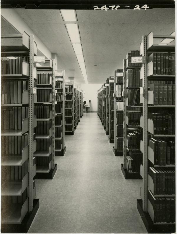 Library stacks with a student working at a desk in the background, ca. 1964