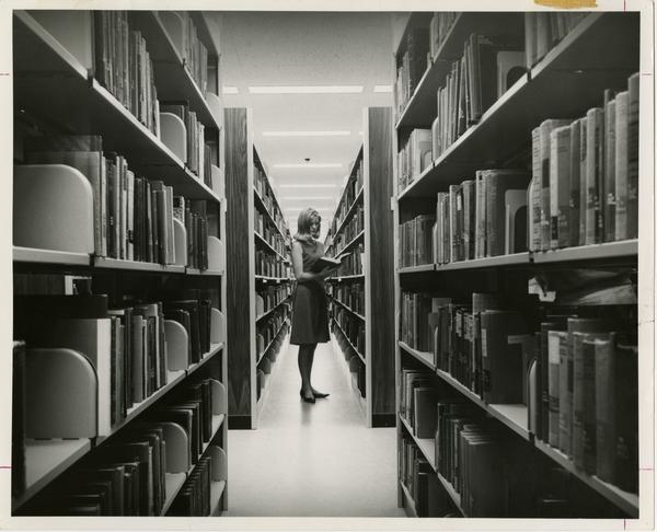 Woman standing between two library stacks examining a book in the University Research Library, ca. 1964