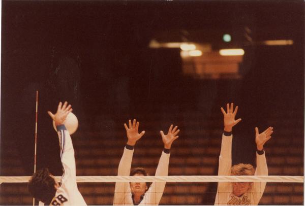 UCLA volleyball player hitting the ball over the net with opposing teammembers attempting to block during a game, 1983