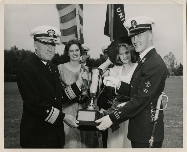 Read Admiral Wilder D. Baker, USN, presents the winner's cup to Sherwood L. Simpson, Naval R.O.T.C. cadet with color girls, Jayne and Molly Cosgrove in the background.