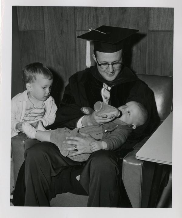 Graduate of the medical school feeds his child as another child looks on after the ceremony, 1956