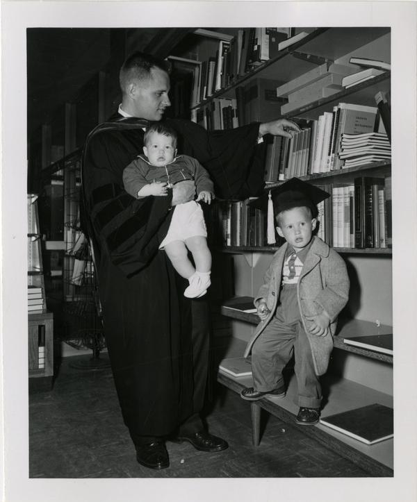 Graduate student of the medical school holding his child while his other child wears his graduation cap in the library after the ceremony, 1956