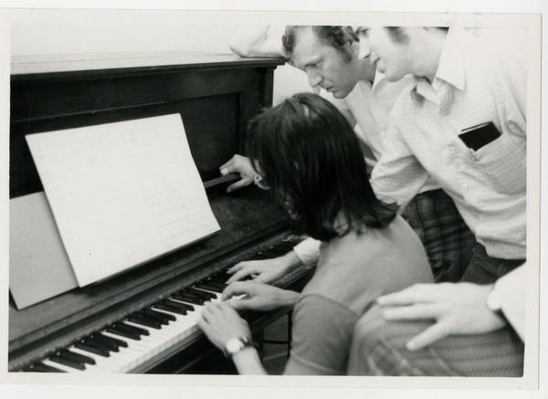 Student plays at the piano while instructors look over her shoulder and guide her, 1972