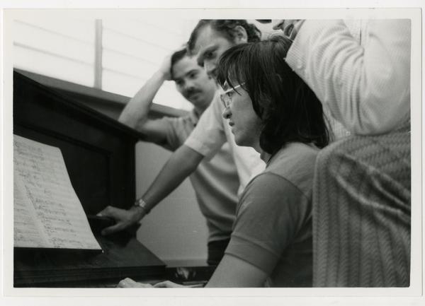 Student sits at the piano while instructors look over her shoulder during composition class, 1972