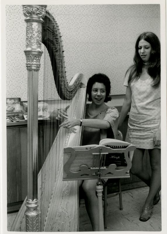 Student strums the harp while another student looks on in the practice room, 1972