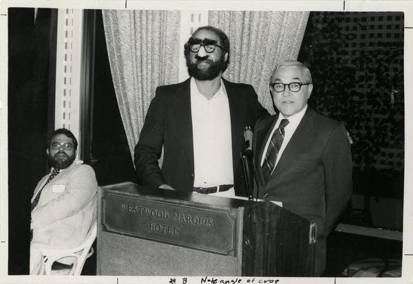 Two unidentified males behind a Westwood Marquis Hotel podium while a third male sitting in a chair