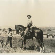 Man on horse and man standing on a grassy hill with farmland in the background