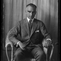 Portrait of Adelbert Bartlett, in suit and tie, seated in wicker chair in front of curtain, [1923?]