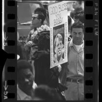 Woman in crowd holding sign on which is written "God Bless You Bobby…Rest In Peace" surrounding a picture of Robert F. Kennedy, 1968