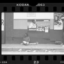 Man sitting on skid row sidewalk below cigarette ads in Los Angeles, Calif., 1986